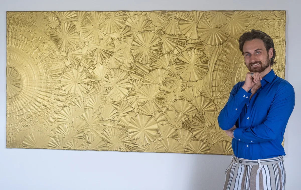 Der Künstler arbeitet gerne mit Strukturpaste und ausdrucksstarken Farben. Man sieht einen Mann, der neben einem goldenen Bild steht.